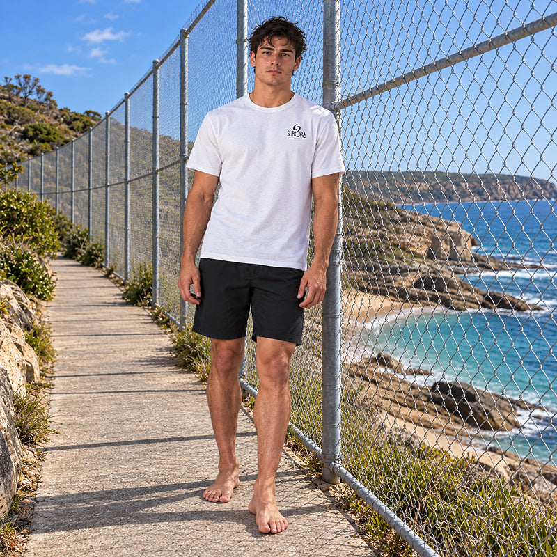 Front view of the Subora Board Meeting Tee in White on a male model walking a coastal clifftop path, showing the black Subora logo chest print