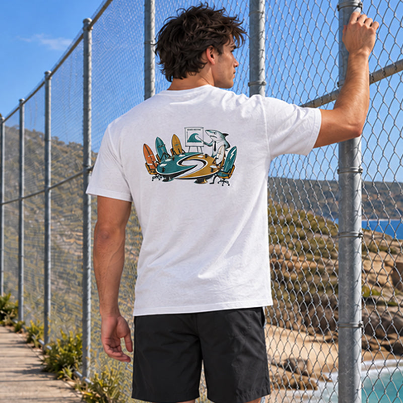 Back view of the Subora Board Meeting Tee in White on a male model at a clifftop fence, showing the illustrated back print of surfboards in ties around a Subora S table with a shark presenting a wave chart