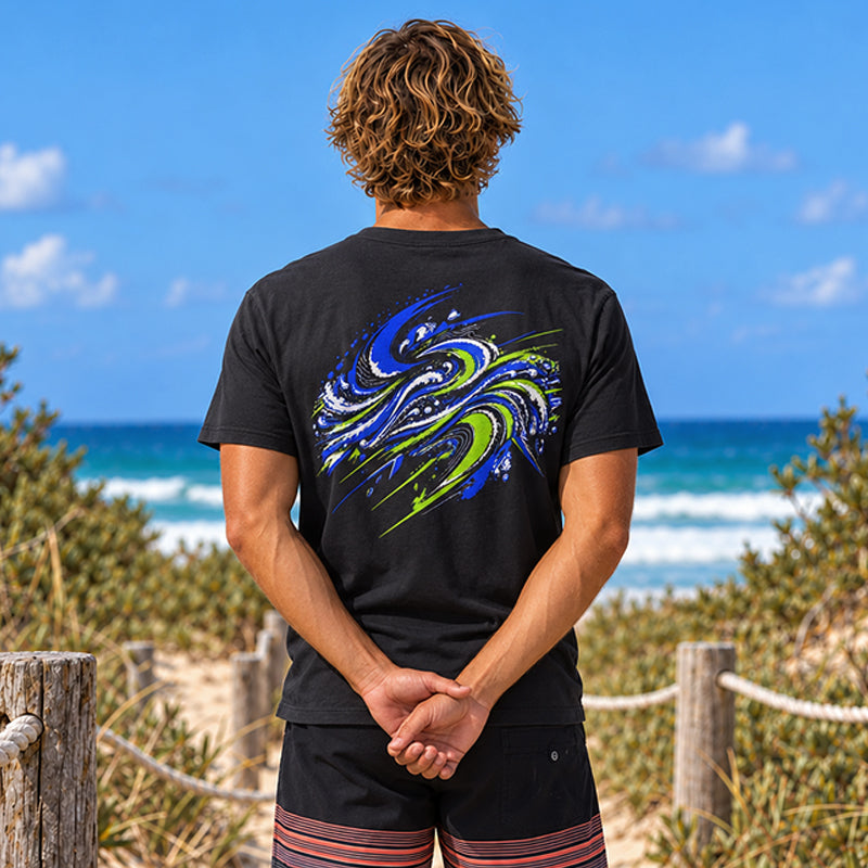 Back view of the Subora Motion Tee in Black on a male model standing on a beach dune track, showing the full-colour blue and lime green swirling wave back print
