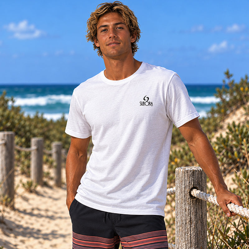 Front view of the Subora Motion Tee in White on a male model leaning on a beach fence, showing the black Subora logo chest print