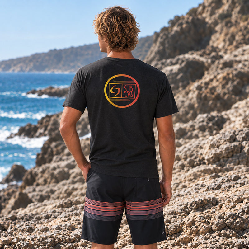 Back view of the Subora Rainbow Logo Tee in Black on a male model standing on coastal rocks, showing the rainbow gradient circle logo back print