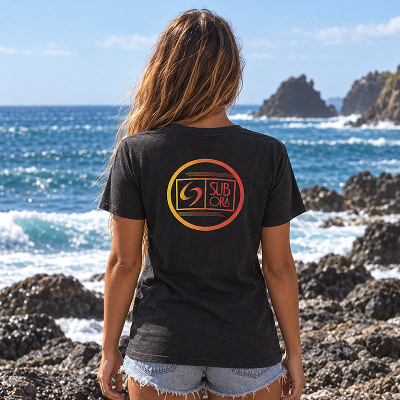 Back view of the Subora Rainbow Logo Tee in Black on a female model standing on coastal rocks, showing the rainbow gradient circle logo back print