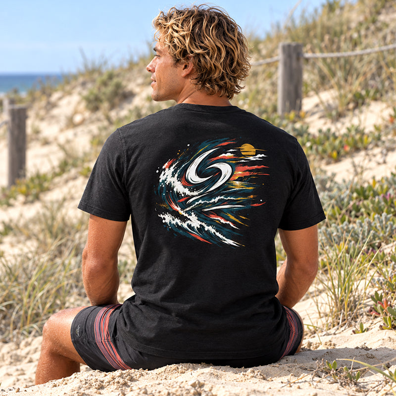 Back view of the Subora Sunset Tee in Black on a male model sitting on beach dunes, showing the full-colour swirling sunset wave back print