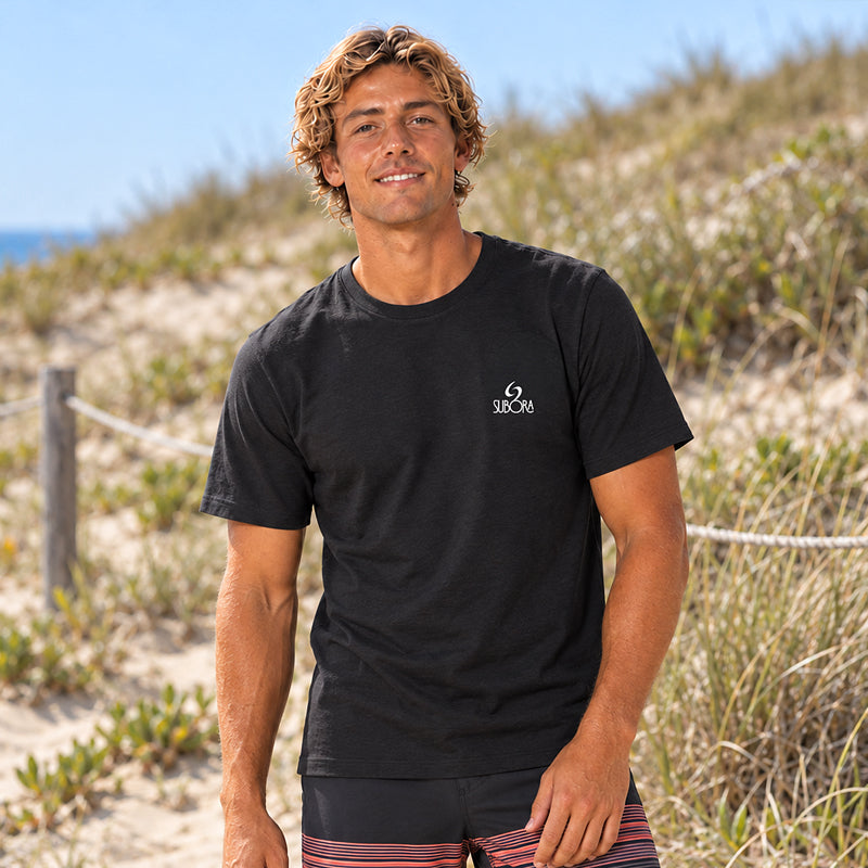 Back view of the Subora Sunset Tee in Black on a male model sitting on beach dunes, showing the full-colour swirling sunset wave back print