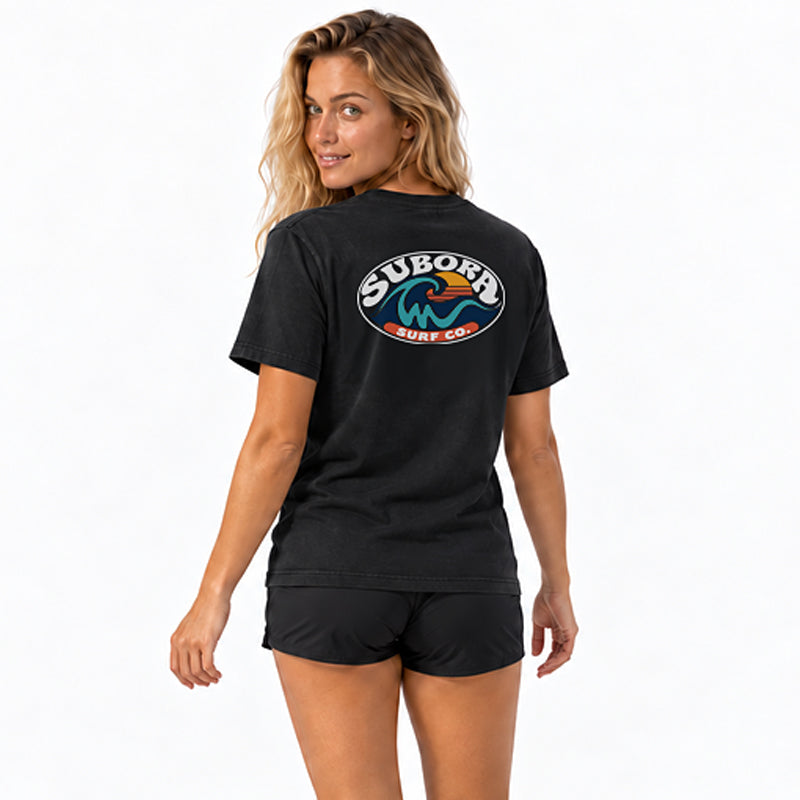 Back view of the Subora Sunwash Tee in Black on a female model, showing the retro oval Subora Surf Co. badge back print with a teal wave, orange setting sun and white lettering