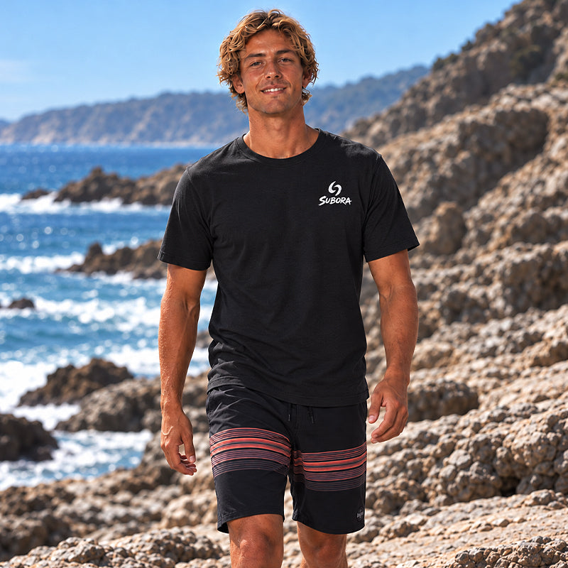 Back view of the Subora Sun, Sand Surf Tee in Black on a male model sitting on coastal rocks, showing the full-colour abstract sun, sand and wave back print