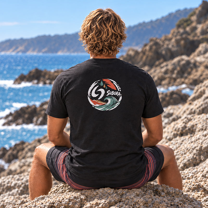 Back view of the Subora Sun, Sand Surf Tee in Black on a male model sitting on coastal rocks, showing the full-colour abstract sun, sand and wave back print